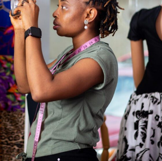 A Nigerian female fashion marketing entrepreneur using a smartphone to create marketing content in a clothing studio beside a mannequin wearing a black and white dress.