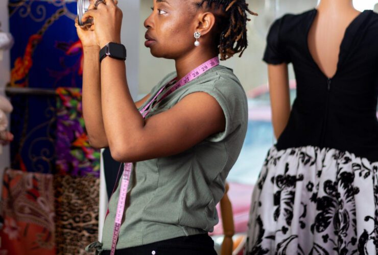 A Nigerian female fashion marketing entrepreneur using a smartphone to create marketing content in a clothing studio beside a mannequin wearing a black and white dress.