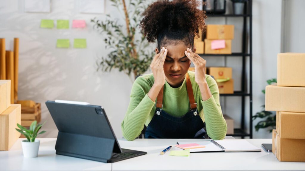 A frustrated female entrepreneur sitting at a desk with packaging boxes, a tablet, and business notes while holding her head in stress.