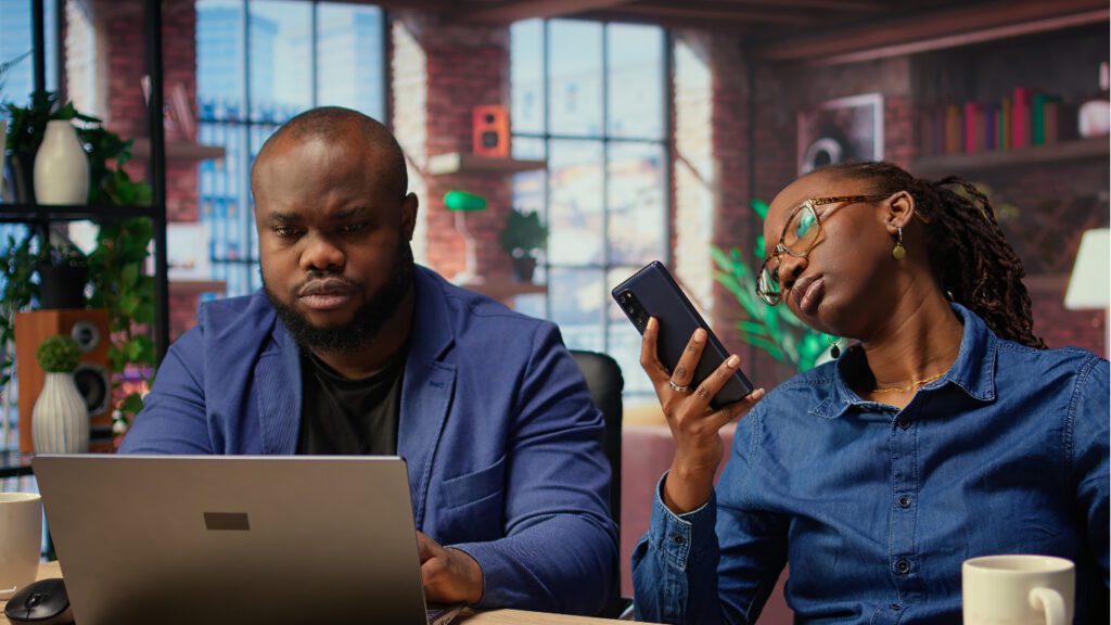 A man in a blue blazer focused on his laptop while a woman beside him scrolls through her smartphone with a skeptical expression — representing how consumers research and silently judge businesses online before making decisions.