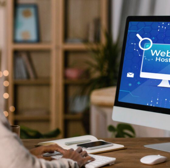 A person sitting at a desk working on a desktop computer displaying a blue "Website Hosting" graphic with icons for cloud storage, security, search, and HTTPS representing the process of setting up and managing website hosting for a business online presence