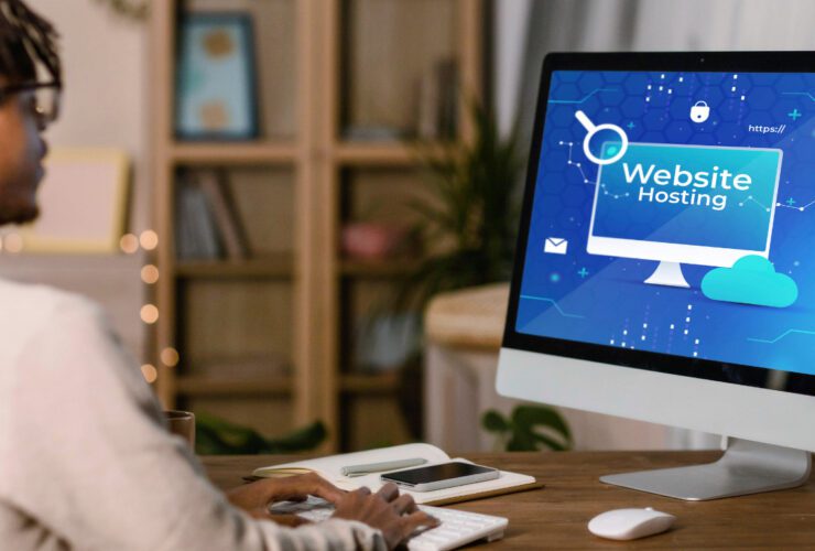 A person sitting at a desk working on a desktop computer displaying a blue "Website Hosting" graphic with icons for cloud storage, security, search, and HTTPS representing the process of setting up and managing website hosting for a business online presence