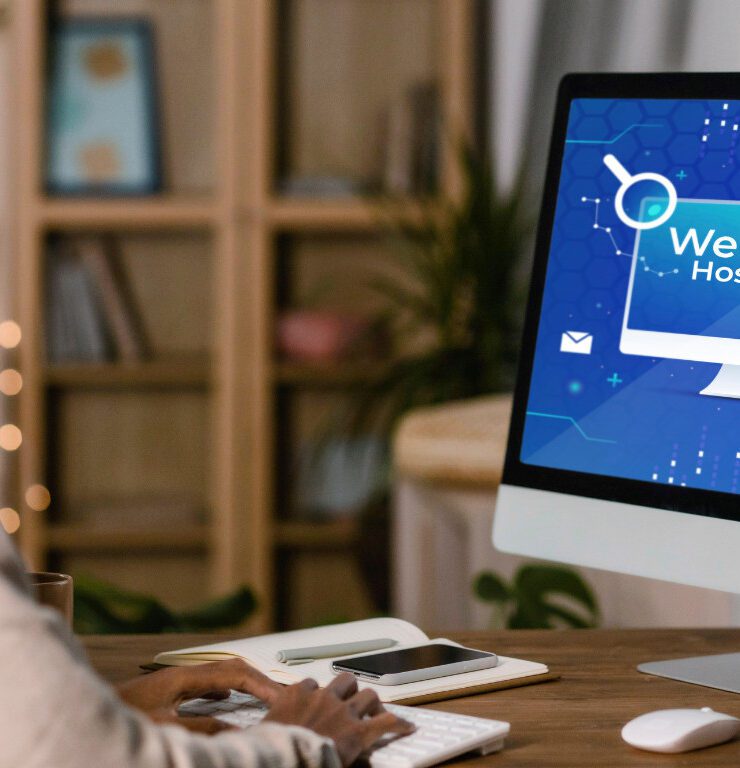 A person sitting at a desk working on a desktop computer displaying a blue "Website Hosting" graphic with icons for cloud storage, security, search, and HTTPS representing the process of setting up and managing website hosting for a business online presence