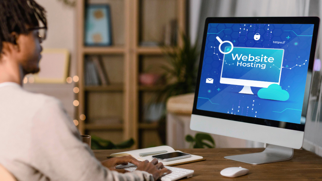 A person sitting at a desk working on a desktop computer displaying a blue "Website Hosting" graphic with icons for cloud storage, security, search, and HTTPS representing the process of setting up and managing website hosting for a business online presence