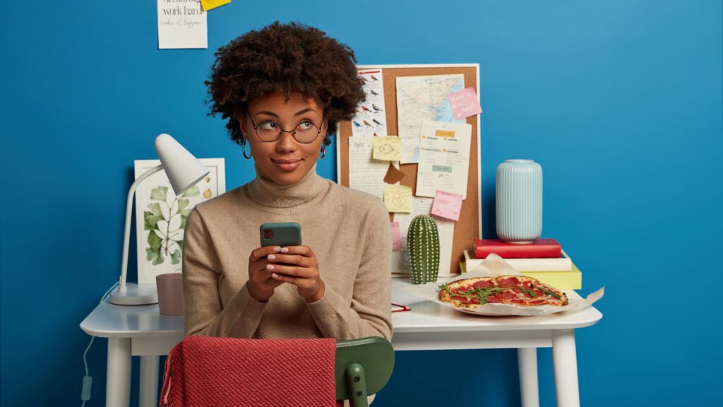 A young woman with natural hair and glasses sits in front of a busy vision board, holding her smartphone and glancing sideways, representing the everyday consumer who discovers and engages with businesses through social media.