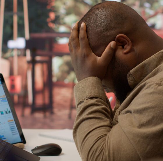 A frustrated business professional is sitting at a desk with a laptop and tablet displaying analytics social media dashboards, holding his head in his hands symbolizing the overwhelm of managing business growth without a clear digital marketing strategy.