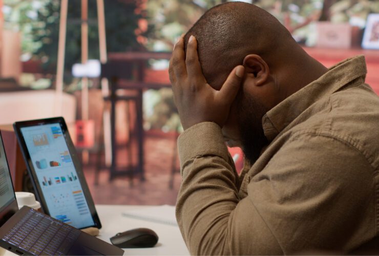 A frustrated business professional is sitting at a desk with a laptop and tablet displaying analytics social media dashboards, holding his head in his hands symbolizing the overwhelm of managing business growth without a clear digital marketing strategy.