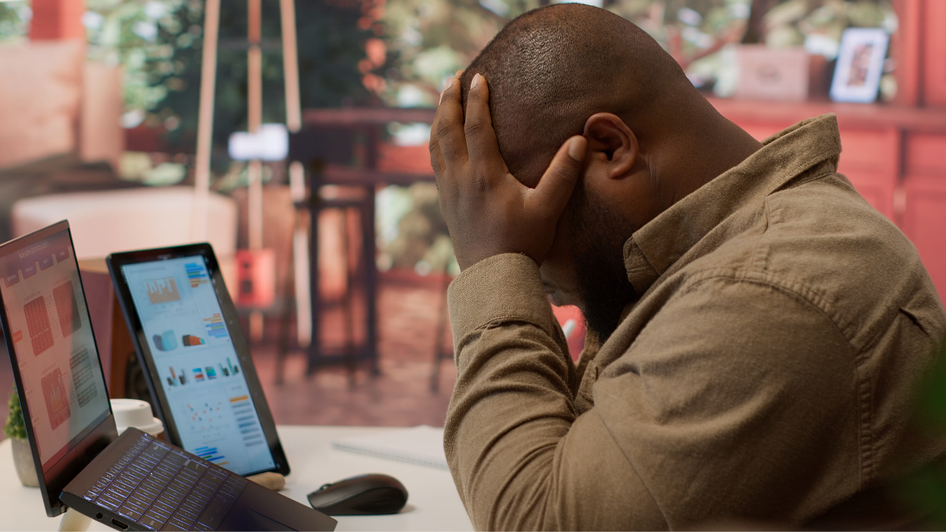 A frustrated business professional is sitting at a desk with a laptop and tablet displaying analytics social media dashboards, holding his head in his hands symbolizing the overwhelm of managing business growth without a clear digital marketing strategy.